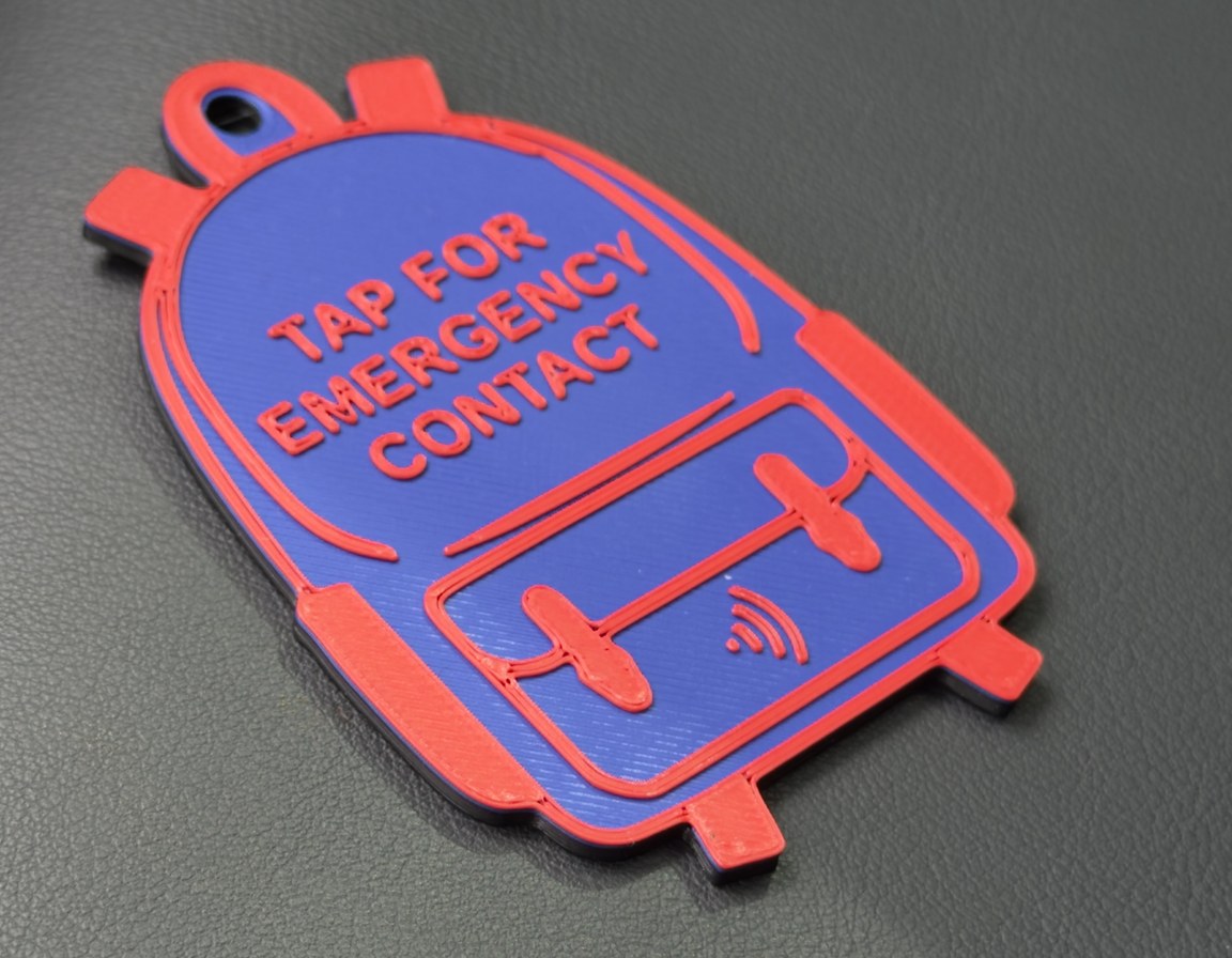 The backpack tag at a three-quarter angle on dark textured leather, TAP FOR EMERGENCY CONTACT raised in coral-red across the blue flap