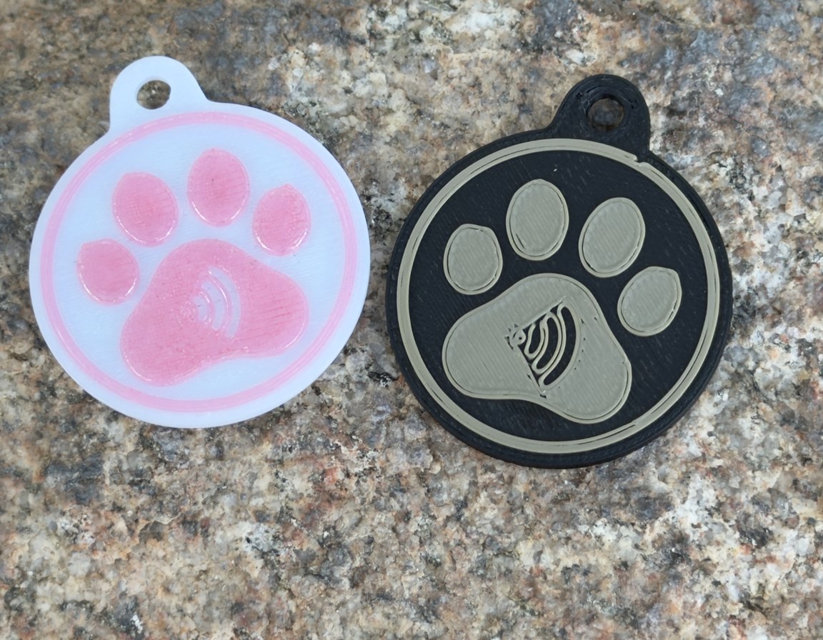 Both Replegend paw tags side by side on speckled granite: a white tag with the paw sunk into pink on the left, a black tag with the paw raised in tan on the right